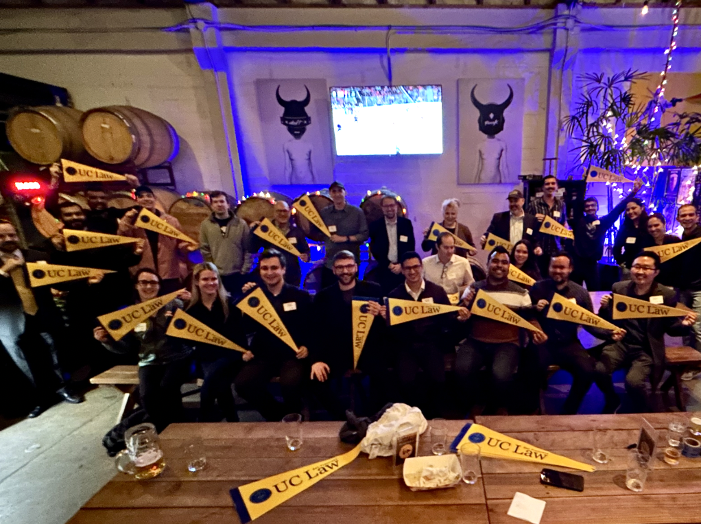 A group of UC Law alumni poses together at a festive event, holding banners with "UC Law" written on them in a brewery setting.